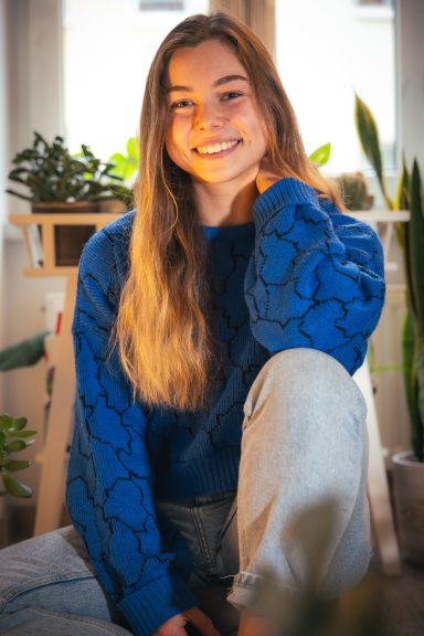 Lächelnde junge Frau mit langen Haaren, blauer Pullover, sitzt in einem hellen Raum.