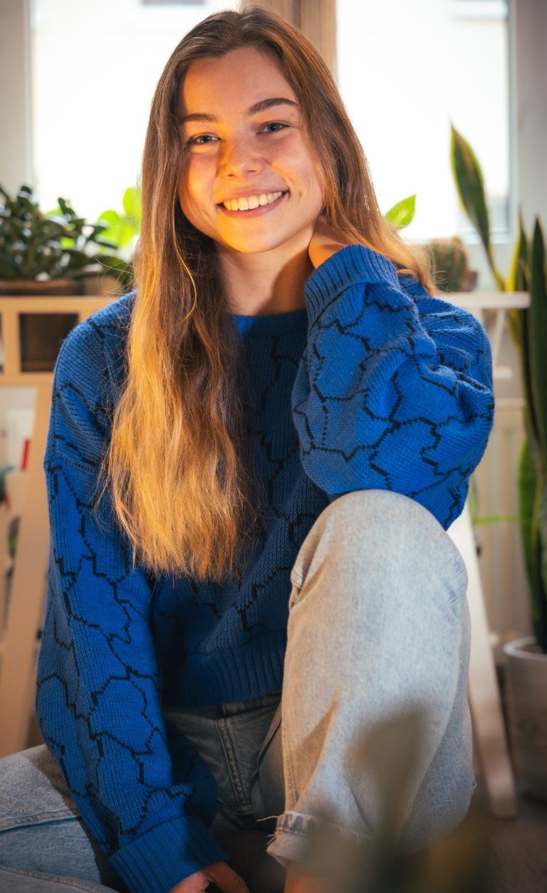 Das bin ich: Emi von emi.atmet Junge Frau mit langen, braunen Haaren in einem blauen Pullover, lächelt freundlich.